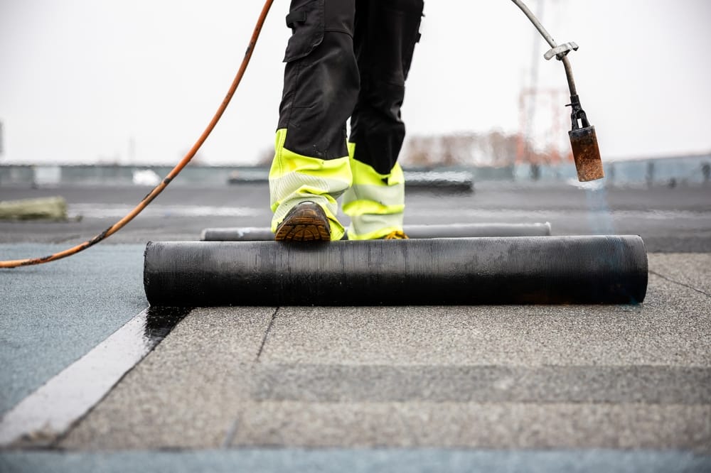 Couvreur professionnel appliquant du goudron au chalumeau sur une nouvelle membrane de toiture pour l'étanchéité d'un toit plat, chantier avec équipement de sécurité.