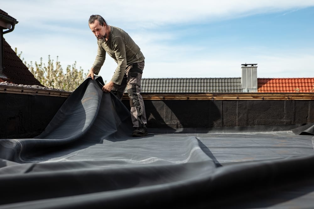 Un artisan déroule une bâche en caoutchouc épais sur un toit plat pour refaire l'étanchéité et réparer le revêtement de bitume endommagé.