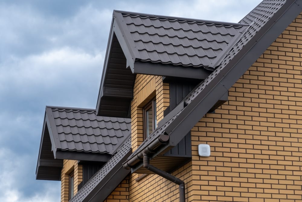 Exterior view of a modern home with a metal roof, brick wall, and drainpipe against a cloudy sky.