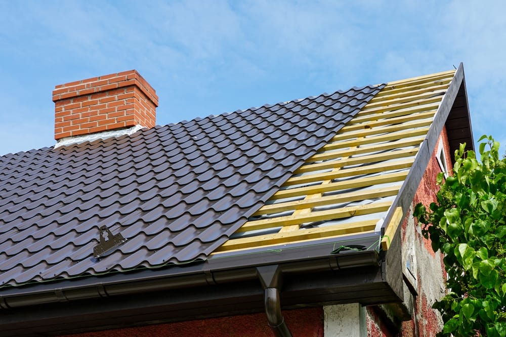 Vue rapprochée d’une installation de toiture en cours avec des tuiles métalliques et une cheminée sur une maison, montrant les liteaux en bois.