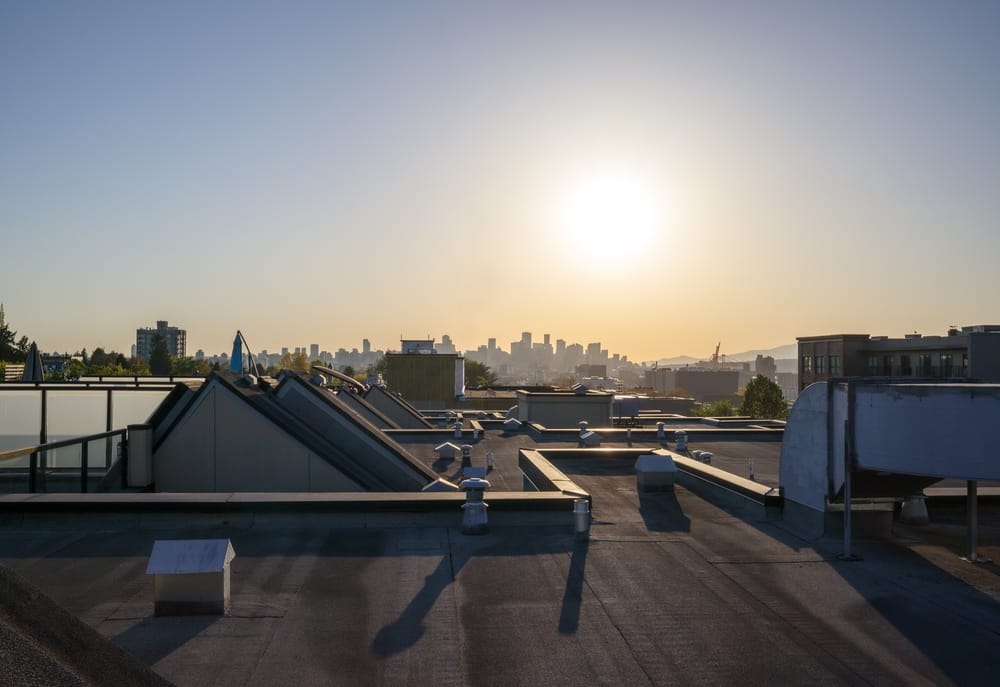 Flat roof with city skyline during sunset. Backlit view of downtown Vancouver far back. 2-ply SBS or modified bitumen roofing system with roof vents, hvac unit and roof hatches. Selective focus.