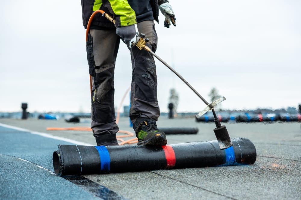 Un travailleur en équipement de protection utilise une torche au propane pour appliquer une membrane de toiture noire sur un toit plat, avec des matériaux supplémentaires et un ciel nuageux visible.
